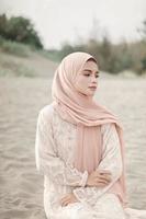 schönes islamisches weibliches modell, das hijab-mode trägt, ein modernes hochzeitskleid für muslimische frau, die im sand und am strand sitzt. Porträt eines asiatischen Mädchenmodells mit Hijab, das sich am Strand mit Bäumen amüsiert foto