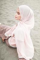 schönes islamisches weibliches modell, das hijab-mode trägt, ein modernes hochzeitskleid für muslimische frau, die im sand und am strand sitzt. Porträt eines asiatischen Mädchenmodells mit Hijab, das sich am Strand mit Bäumen amüsiert foto