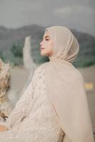 schönes islamisches weibliches modell, das hijab-mode trägt, ein modernes hochzeitskleid für muslimische frau, die im sand und am strand sitzt. Porträt eines asiatischen Mädchenmodells mit Hijab, das sich am Strand mit Bäumen amüsiert