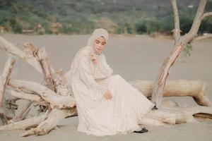schönes islamisches weibliches modell, das hijab-mode trägt, ein modernes hochzeitskleid für muslimische frau, die im sand und am strand sitzt. Porträt eines asiatischen Mädchenmodells mit Hijab, das sich am Strand mit Bäumen amüsiert