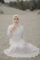 schönes islamisches weibliches modell, das hijab-mode trägt, ein modernes hochzeitskleid für muslimische frau, die im sand und am strand sitzt. Porträt eines asiatischen Mädchenmodells mit Hijab, das sich am Strand mit Bäumen amüsiert foto