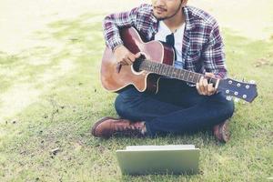 junger Hipster-Mann übte Gitarre mit Laptop im Park, glücklich und spielte gerne Gitarre. foto