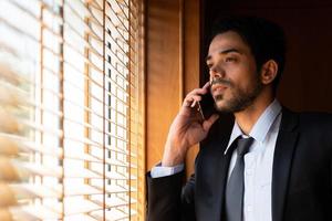 Porträt eines jungen Geschäftsmannes, der aus dem Fenster schaut und das Smartphone verwendet, um mit dem Kunden zu sprechen. Geschäftskonzept foto