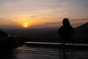Silhouette einer Frau, die auf der Terrasse sitzt und den Sonnenuntergang über dem Berg betrachtet foto