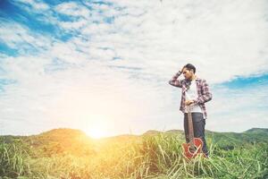 Junger Hipster-Mann, der eine Gitarre mit einem Spaziergang in der Natur hält und sich an einem sonnigen blauen Himmelstag auf dem Feld entspannt. foto