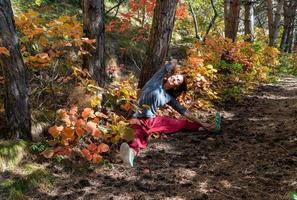 lächelnde Frau mit roter indischer Hose, die im Wald unter den Bäumen auf dem Weg mit Herbstlaub sitzt, Yoga-Posen foto