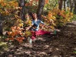 lächelnde Frau mit roter indischer Hose, die im Wald unter den Bäumen auf dem Weg mit Herbstlaub sitzt. Porträt einer glücklichen Frau foto