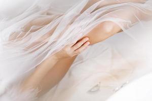 Morgen der Braut. Zugeschnittenes Foto einer schönen jungen Frau trägt einen weißen langen Schleier, einen Bademantel und Unterwäsche, die lachend sitzen und Spaß im Bad auf weißem Hintergrund haben, entspannen. Hochzeitstag.