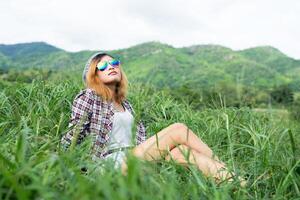 Schöne Hipster-Frau, die auf einer Wiese mit Natur und Bergen im Hintergrund sitzt. foto