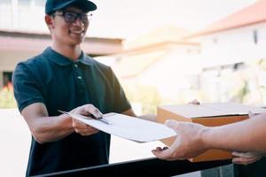 junger asiatischer mann lächelt, während er der frau, die ein dokument hält, einen pappkarton liefert, um unterschrift zu unterzeichnen. foto