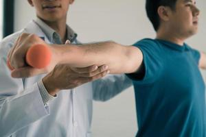 asiatischer junger männlicher physiotherapeut, der patienten beim heben von hanteln hilft, übungen im büro. foto