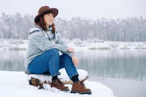 Frau mit braunem Hut und Pullover, die am Ufer eines schneebedeckten Sees und Waldes sitzt foto