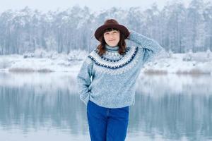 Eine lächelnde Frau mit braunem Hut und Pullover steht am Ufer eines schneebedeckten Sees und Waldes foto