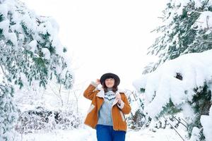 junge frau mit hut und warmer jacke, die zwischen schneebedeckten bäumen steht und den ersten schnee genießt foto