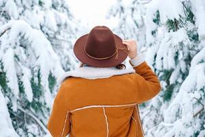 Frau mit Hut und warmer Jacke, die auf einem verschneiten Wald steht. Ansicht von hinten foto