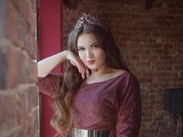 Porträt einer schönen jungen, hübschen, süßen Prinzessin in langem lila Kleid und Krone der Königin, mit langen Haaren und Make-up im minimalistischen Loft-Interieur, das am Fenster steht foto