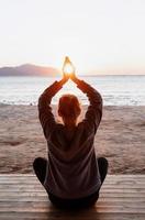 Junge gesunde Frau, die Yoga praktiziert und Händchen hält, in Meditationspose mit Sonne zwischen ihnen am Strand bei Sonnenaufgang foto