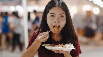 asiatische junge frau reist in bangkok, thailand, schöne weibliche fühlen sich glücklich beim gehen und essen pad thai an der khao san road. frauen reisen essen straßenessen im thailand-konzept. foto