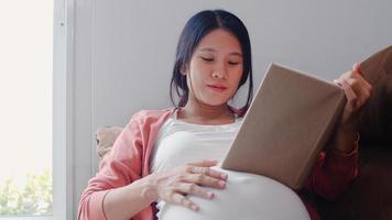 junge asiatische schwangere frau las ein buch für baby im bauch. mutter fühlt sich glücklich lächelnd positiv und friedlich, während sie auf das kind liegt, das auf dem sofa im wohnzimmer zu hause konzept liegt. foto