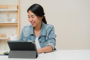 junge asiatische frau, die mit einem tablet arbeitet, soziale medien überprüft und kaffee trinkt, während sie sich zu hause auf dem schreibtisch im wohnzimmer entspannt. zeit zu hause genießen. foto