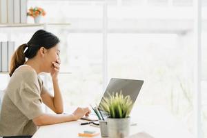 schöne junge lächelnde asiatische Frau, die Laptop auf Schreibtisch im Wohnzimmer zu Hause arbeitet. asiatische Geschäftsfrau, die Notizbuchdokumentfinanzierung und -rechner im Heimbüro schreibt. Zeit zu Hause Konzept genießen. foto