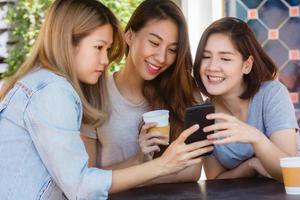 fröhliche asiatische junge frauen sitzen im café, trinken kaffee mit freunden und reden miteinander. attraktive asiatische frau, die kaffee genießt, während sie das smartphone zum sprechen, lesen und texten verwendet. foto