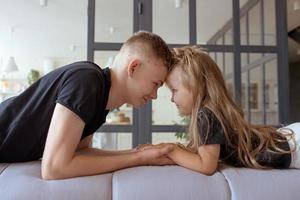 kaukasische geschwister - teenagerjungenbruder und kleine mädchenschwester, die auf einer couch im modernen loftinnenraum sitzen foto