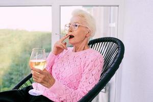 altmodische ältere stilvolle Frau, die auf der Terrasse sitzt und eine Zigarette mit einem Glas Weißwein raucht. schlechte Angewohnheit, Suchtkonzept foto