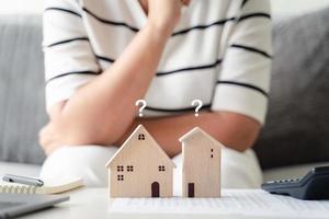 Die Frau entscheidet sich für ein Haus mit einem kleinen Holzhausmodell mit Fragezeichen und plant, eine Immobilie zu kaufen. wählen Sie, was das Beste ist. Konzepte für Wohnungsbaudarlehen. foto