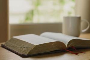 Nahaufnahme der offenen Bibel mit einer Tasse Kaffee für die Morgenandacht auf einem Holztisch mit Fensterlicht. christlicher Hintergrund foto