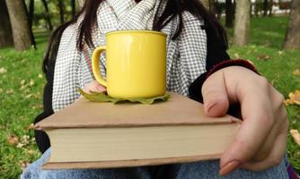 Ein Mädchen in Mantel und Schal im Herbstwald hält an einem warmen Tag in einem Stadtpark ein Buch und eine Tasse mit einem heißen Getränk in den Händen. das Konzept des Lesens, der Entspannung und des Komforts. foto