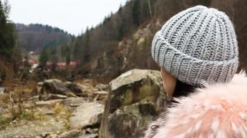 Rückansicht einer Bergtouristin in einer rosafarbenen Jacke mit Pelz und einer grauen Strickmütze vor dem Hintergrund der Berge mit Blick in die Herbstlandschaft des Waldes und des bewölkten Himmels. foto