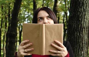 Porträt einer jungen Frau, die ein aufgeschlagenes Buch ohne Titel vor ihr Gesicht hält. Brünette, die an einem sonnigen Tag im Park die Hälfte ihres Gesichts mit einem Buch bedeckt. bildungs- und menschenkonzept, selektiver fokus. foto