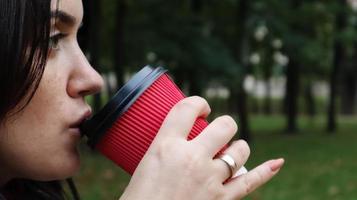 junge stilvolle frau trinkt morgens heißen kaffee in rotem öko-pappbecher draußen im herbstpark. Nahaufnahme Seitenansicht der jungen Frau mit Kaffeetasse zum Mitnehmen. foto