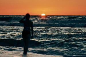 weibliche Silhouette in blauen Meereswellen bei Sommersonnenuntergang, halbe Sonne unter Horizont, Strandurlaub foto