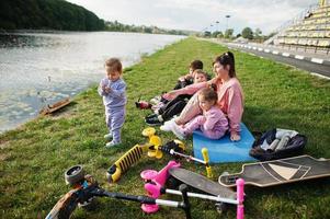 junge stilvolle mutter mit vier kindern, die auf gras gegen see sitzen. Sportfamilie verbringt ihre Freizeit im Freien mit Rollern und Schlittschuhen. foto