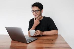 junger asiatischer mann lächelt und genießt es, wenn er zu hause mit laptop und einer tasse kaffee arbeitet. Ein Mann sitzt auf dem Bett und arbeitet. Arbeit von zu Hause aus Konzept foto