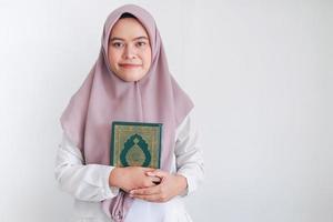 junge asiatische islamfrau mit kopftuch hält den heiligen al quran mit einem lächeln und einem glücklichen gesicht in der hand. indonesische Frau auf grauem Hintergrund foto