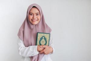 junge asiatische islamfrau mit kopftuch hält den heiligen al quran mit einem lächeln und einem glücklichen gesicht in der hand. indonesische Frau auf grauem Hintergrund foto