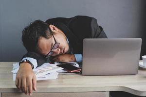asiatischer geschäftsmann, der am schreibtisch mit geschlossenen augen auf grauem hintergrund einschläft, überarbeiteter junger mann, unmotivierter arbeiter, der am arbeitsplatz schläft, langweilige routinearbeit foto
