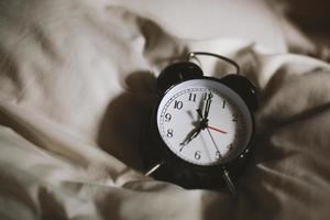 Klassischer schwarzer Wecker, der sieben Uhr mit einem Bett im Hintergrund zeigt und morgens aufwacht foto