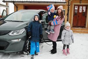 Junge isländische Mutter mit Kindern hält im Winter Islandfahnen und lädt Elektroautos im Hof ihres Hauses auf. foto