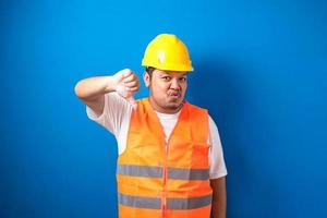 Junger dicker asiatischer Bauarbeiter mit orangefarbener Sicherheitsweste und Helm, der unglücklich und wütend aussieht foto