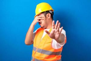 Fetter asiatischer Bauarbeiter in Uniform und Helm über isoliertem blauem Hintergrund, der die Augen mit der Hand bedeckt foto