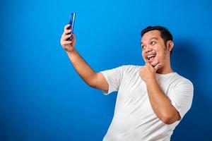 Studioporträt eines jungen ostasiatischen Mannes in weißem T-Shirt, der einen Videoanruf tätigt und ein Selfie vor blauem Hintergrund macht foto