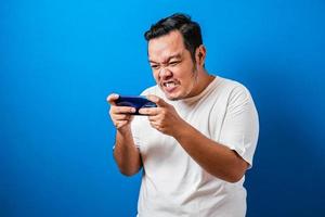Wütender, dicker Asiate, der Spiele auf dem Tablet-Smartphone vor blauem Hintergrund spielt foto