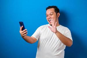 Studioporträt eines jungen ostasiatischen Mannes in weißem T-Shirt, der einen Videoanruf tätigt und ein Selfie vor blauem Hintergrund macht foto