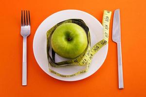 grüner Apfel auf Teller mit Maßband, Messer und Gabel auf orangefarbenem Hintergrund foto