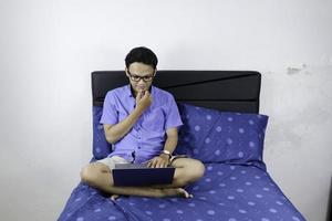 junger asiatischer mann lächelt und genießt es, wenn er zu hause arbeitet. Ein Mann, der auf dem Bett sitzt und mit einem Laptop arbeitet. Arbeit von zu Hause aus Konzept. foto