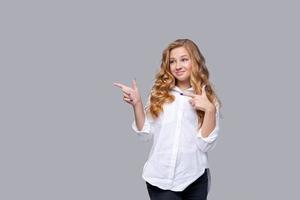 Zuverlässiges, fröhliches Mädchen im weißen Hemd zeigt mit dem Finger auf eine leere Stelle foto